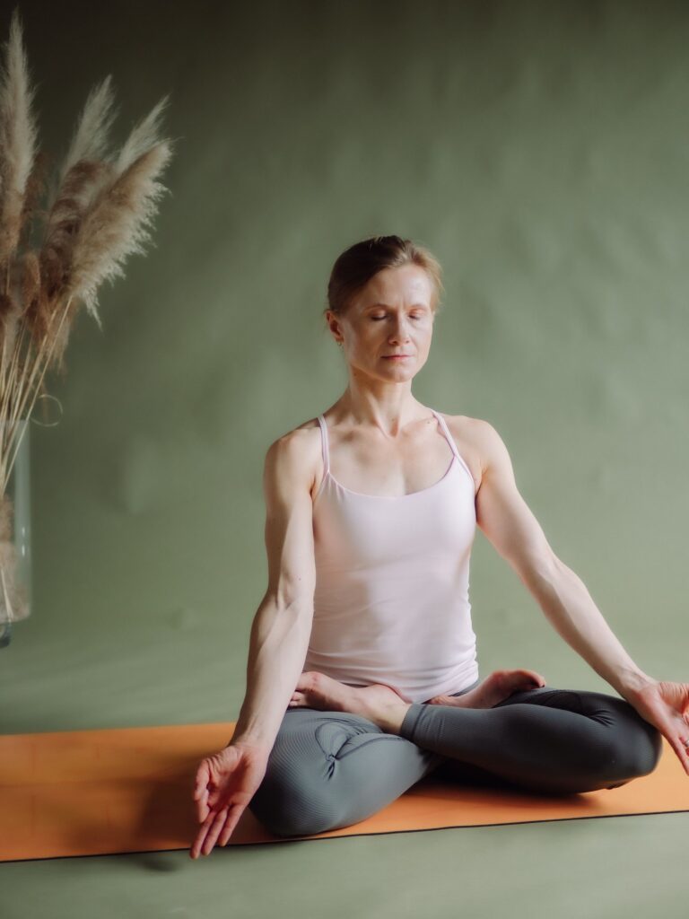 Yogalehrerin Ela Buchholz – Meditationssitz mit Chin Mudra vor gruener Wand, Pampasgras, warmes Licht, Hochformat